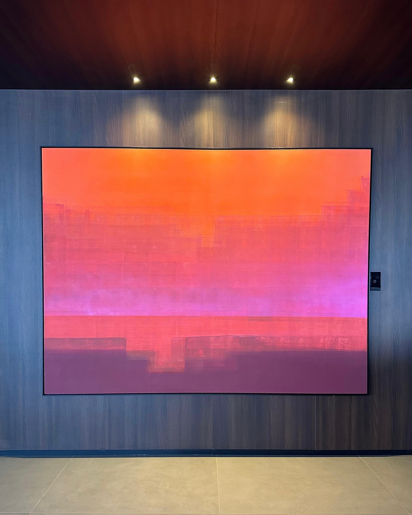 Abstract painting in a gradient of orange, pink and purple, displayed on a dark wooden wall under spotlights